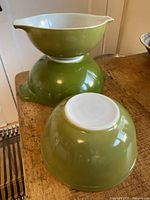 Three avocado green Pyrex mixing bowls stacked and separately placed on wooden table, showing handles and spouts.