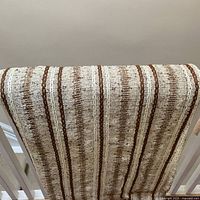 Close view of textured loosely woven drapery panel showing vertical chocolate and off-white yarn stripes on a creamy background, draped over railing.