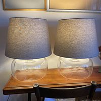 Pair of clear glass table lamps with gray textured shades placed on a wooden surface.