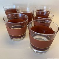 Array of six clear glass lowball glasses with horizontal burgundy and cinnamon stripes bordered by thin gold bands shown from a side angle.
