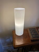 Photo of tubular frosted white glass lamp illuminated on a wooden table against a plain wall.