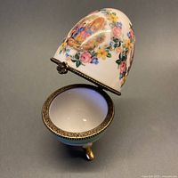 View of vintage hand painted hinged Limoges egg with antique scene and floral wreath on upper half, open to show interior and brass hinge