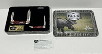 Top-down view of the tin box containing two Buck combo folding pocket knives, black foam lining, and a Buck/Realtree card.