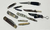 Top view showing all knives and pocket cutters laid out on a white surface, displaying a variety of sizes and handles including folding knives and fixed blades with sheaths.