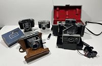 Overview of four cameras and accessories on white surface: brown leather-case folding camera, small silver camera, Ansco box, two bellows-type cameras including Polaroid