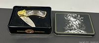 Wildlife Collection Bass folding box cutter with open blade displayed inside original black tin featuring bass fish illustration.
