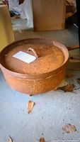 Rusty cast iron pot with lid and loop handle sitting on a concrete floor with cardboard in background