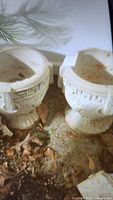 Two round cement planters with geometric relief designs shown side by side on outdoor ground with dirt and leaves.