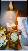 Photo showing the Coca-Cola solid wood framed clock leaning against the bed and the crystal lamp on a bedside table with the lamp lit.