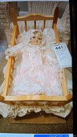 Photo showing the baby doll dressed in a pink lace gown and bonnet lying in the wooden cradle with cream ruffled bedding.