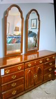 Front view of solid wood dresser with two attached mirrors, brass hardware, in a room setting showing scratches on surface.