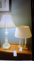 Two table lamps on wooden surface, one brass base with dual bulbs and pleated shade, the other glass and brass base with tapered shade.