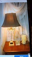 Photo showing a table lamp with dark brown shade, one candle in a wire holder, and a Mikasa crystal clock on a wooden surface in front of a window with white curtains.