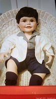 Close-up of Baby Donnie doll sitting in a wicker chair wearing blue checked shirt, white jacket, navy shorts, socks, and black shoes