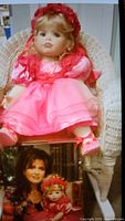 Marie Osmond Friendship collectible doll sitting on a white wicker chair, wearing a pink satin dress and matching hat. The box with image of Marie Osmond holding the doll is visible beneath the doll.