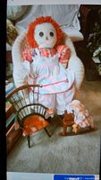 Photo showing a child size white rattan rocker with a large Raggedy Ann style doll seated on it, a small wooden doll chair, and a baby doll on a wood rocking horse on carpeted floor.