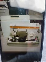 Photo of the Singer portable sewing machine on its base, showing the orange/gold Singer logo strip, rotary dial, foot pedal and needle area.