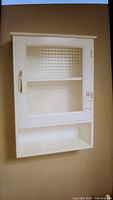 Front view of white wall cabinet showing glass door, handle, and open shelf below.