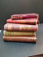 Five antique books stacked and showing spines with titles, colors, and decorative covers.