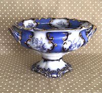Side view of large blue and white flow blue fruit bowl with gold hand painted trim and ornate handles and base on polka dot background.
