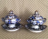 Pair of intricately decorated flow blue sauce tureens with lids and underplates shown side by side on a polka dot surface.