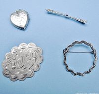 Four sterling silver jewelry items including heart-shaped engraved locket, Victorian bar pin with stones, circular twisted pin, and oval pin on blue background.