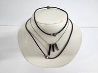 Three necklaces displayed on white neck stand: sterling silver box chain with three geometric bar pendants, black braided leather bracelet, and purple leather necklace with silver clasp.
