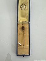 Antique 14K gold stick pin with square-cut red stone and diamond accent displayed in its original box with beige satin lining.