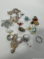 Photo showing a collection of assorted rings, decorative pins and brooches laid out on a flat surface, highlighting varied styles and colors including colorful gemstones, detailed metalwork and enamel accents.
