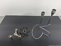 Set of four metal candle holders on black surface: two large loop designs, one smaller starburst candle holder, and one tripod silver holder.