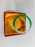 Natural Burmese jade bangle in original red box with yellow satin lining, showing green to white gradient.
