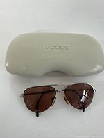 Closed white protective Vogue hard case with embossed logo on top alongside aviator sunglasses with silver metal frames and brown lenses.