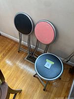 Set of 3 metal collapsing folding stools; two with black padded seats and one with pink seat, showing minor wear and scratches on cushions.