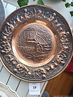 Front view of antique copper wall plate showing embossed watermill house and mountain landscape with ornate floral border.