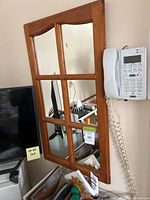 Wall mirror with wooden frame divided into six rectangular panels resembling window panes. Mounted on a wall above some electronics and near a wall phone.