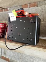 Battery charger unit showing side view and cables coiled on top with copper clamps visible.