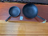 Top-down view of two cast iron pans on wooden surface: a smaller skillet and a larger wok-style pan.