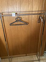Metal clothing rack standing against wood panel wall with several black plastic hangers on it.