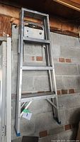 Side view of the aluminum folding step ladder showing steps, shelf, and blue rubber feet against a cinder block wall.