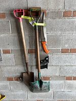 Photo shows two garden shovels with wooden handles and plastic grips, a hoe, an orange pruning saw, and a black hand saw hanging on a concrete wall.