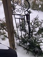 Side view of metal garden trellis standing outdoors in snow, attached loosely to wooden post, displaying scrollwork design and vertical support bars.