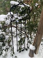 Front view of black metal garden trellis standing outdoors against snowy pine trees, showing overall shape and scrollwork at the top.