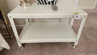 White metal utility table with two rectangular shelves and caster wheels, shown in room setting.