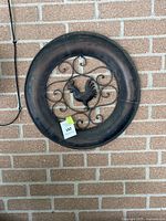 Round metal wall decor with a rooster shape in the center and scroll metalwork, against a brick wall background.