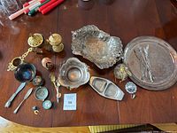 Wide shot showing the full assortment of mixed metal items on a wooden table, including trays, spoons, bowls, and decorative ornaments.