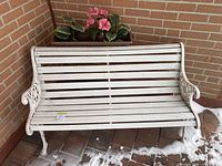 Front view of white outdoor bench with wooden slats and cast metal arms and legs on tiled floor with some snow.