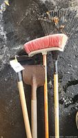 Photo showing four outdoor tools: a metal rake, red-bristle push broom, metal shovel, and metal hoe, all with wooden handles and some dirt on the ground.