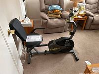 Side view of recumbent bike with padded seat and manual on seat, placed indoors in a living room.