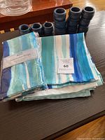 Six cloth napkins with watercolor striped pattern in blues and greens, packaged and loose, displayed with 12 black wooden napkin rings of stacked ring design.