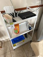 White metal rack on casters with three shelves seen from a front angle showing structure and contents for scale.